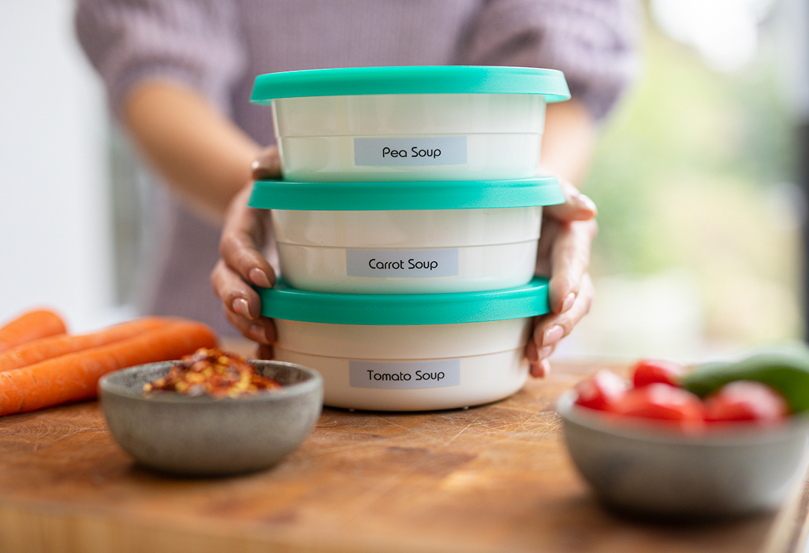 Жена държи три етикетирани кутии Tupperware върху дъска за рязане, върху която има моркови, домати и краставица