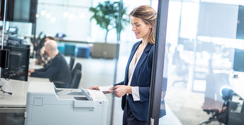Ženska v veliki pisarni uporablja tiskalnik Brother s kartico NFC; PC monitorji, rastline, stoli