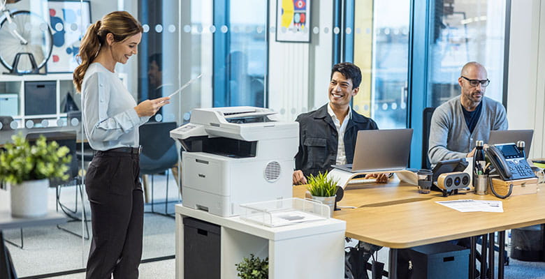 Rušná kancelář, 2 muži sedící a pracující u stolů, žena stojící u tiskárny Brother a držící dokument A4, rostliny, kancelářský nábytek