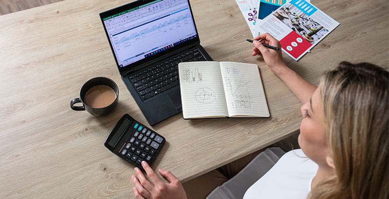 Pohľad zhora na ženu pracujúcu za stolom, na ktorom je zapnutý notebook, zápisník, hrnček s kávou a kalkulačka