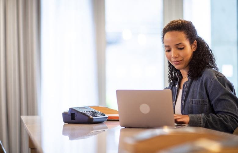 Femeie care stă la birou, cu imprimanta de etichete PT-D410 și laptop