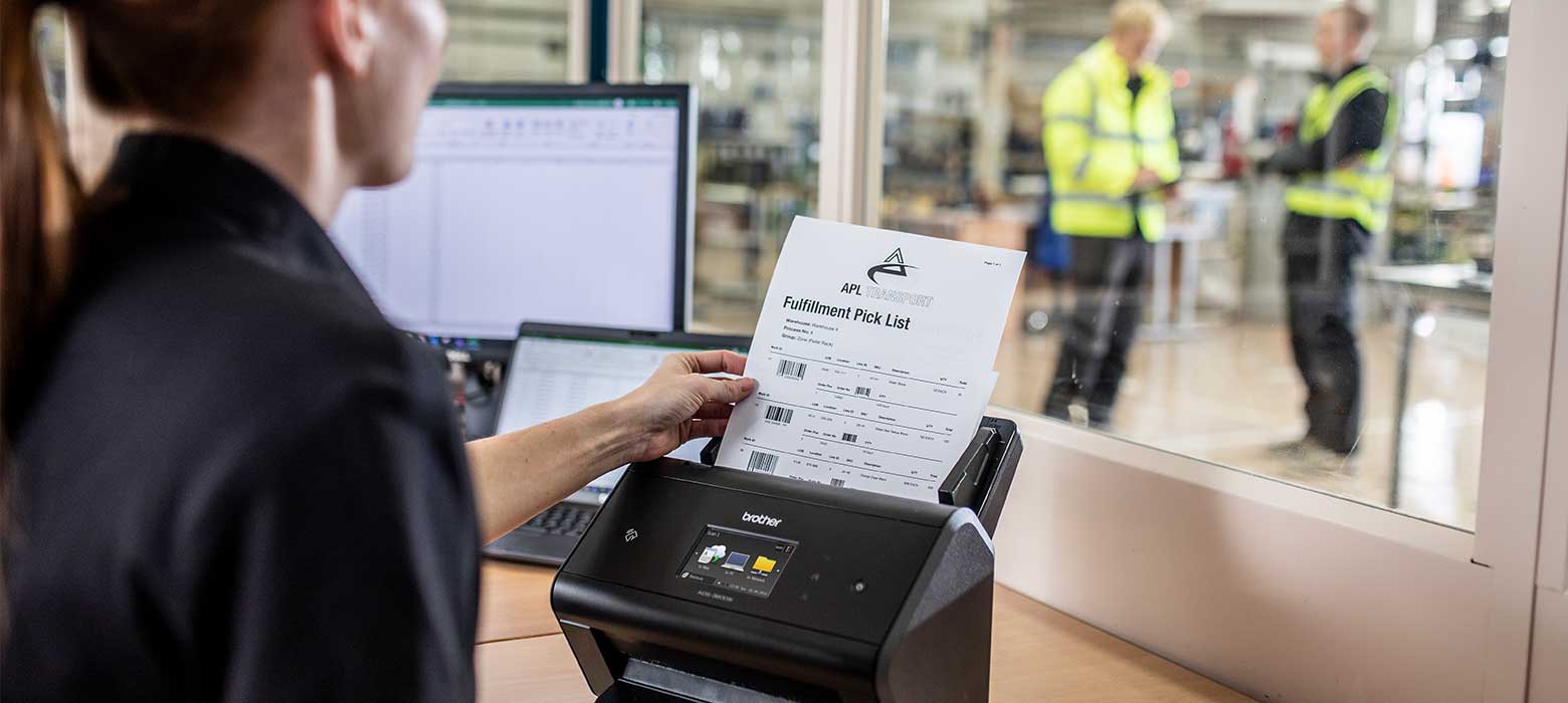 Žena v černém vkládá dokument do skeneru ve skladu s notebookem, monitorem a dalšími pracovníky