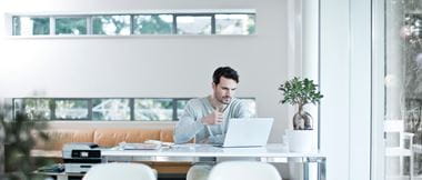 Ein Mann sitzt lächelnd an seinem Küchentisch und arbeitet an einem Laptop. Im Hintergrund sind unscharf ein schwarz-beiger Küchenschrank und ein helles Fenster zu sehen. 