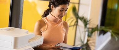 Eine Mitarbeiterin mit orangem Oberteil steht neben einem grossen weissen Bürodrucker und blättert durch eine Broschüre. Im Hintergrund ist unscharf eine Pflanze zu sehen.