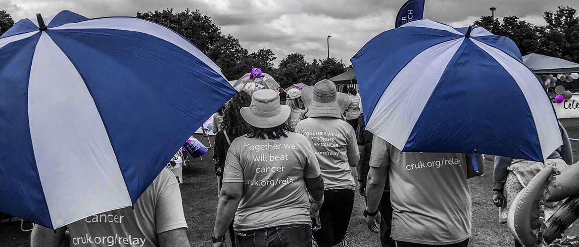 Photo en noir et blanc avec des détails bleus et violets, cette scène montre une marche caritative de Relay for Life sur un circuit de course avec des dizaines de personnes qui aident à lever des fonds pour l’ONG.