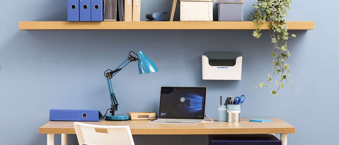Das Bild zeigt eine hybride Arbeitsumgebung mit Laptop, Lampe, Schreibtisch, Dokumenten und Schreibwaren.