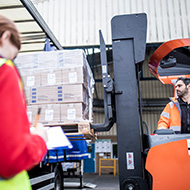 A warehouse employee writing in a clipboard while standing in front of colleague who is sat in a forklift loading cardboard boxes