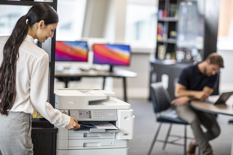 Eine Mitarbeitende nimmt den Ausdruck aus einem Brother Drucker. Im Hintergrund arbeitet ein Mann an einem Laptop