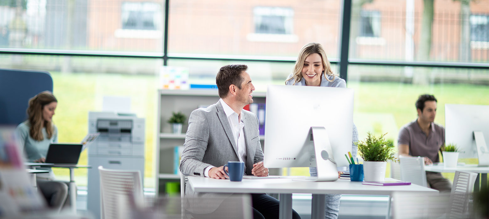 Man sat at desktop computer speaking to woman with long blond hair, people working on laptops in the background, printers, mugs, plants, chairs, office