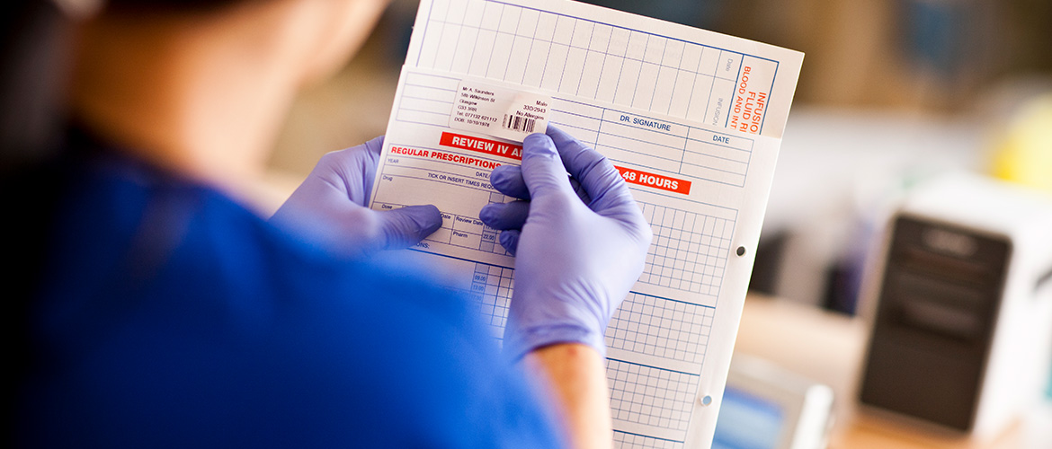 KRANGEPFLEGERIN KLEBT Barcode-Etikett auf Patientenakte. EtikettendRucker im Hintergrund.