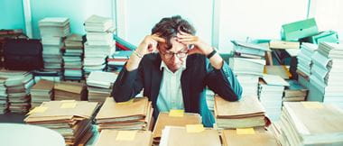 Ein Anwalt hält sich den Kopf, während er an einem Schreibtisch in einem Büro sitzt, umgeben von Stapeln von Aktenordnern und Dokumenten.