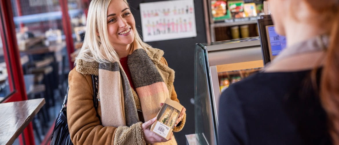  Eine blondhaarige junge Frau, die einen herbstlichen Mantel und einen Schal trägt, kauft in einem Einzelhandelsgeschäft für Feinkost ein vorgefertigtes, deutlich gekennzeichnetes Sandwich.
