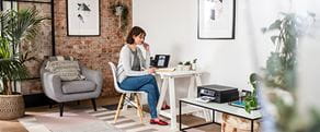 Frau sitzt zu Hause am Schreibtisch und schaut auf ihren Notizblock und Laptop mit Drucker, Pflanzen und einem Stuhl im Vordergrund