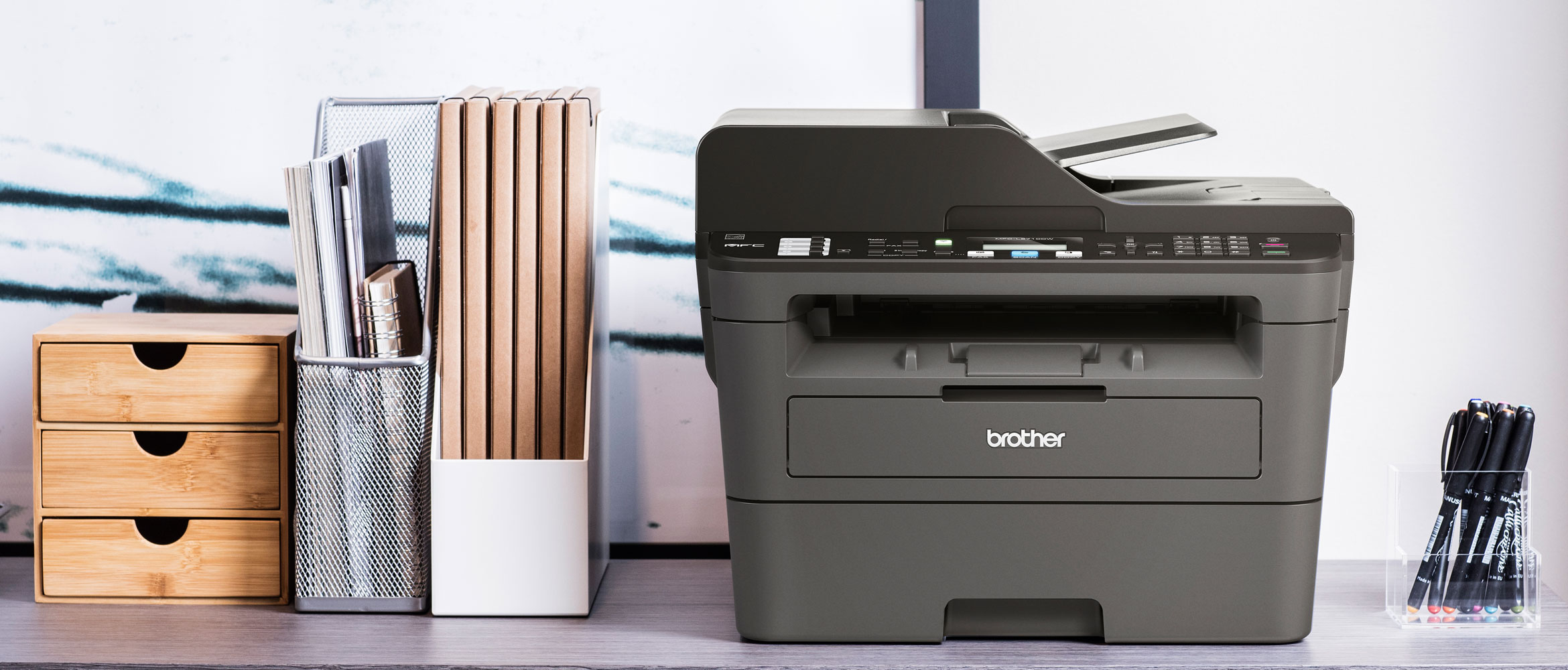 MFC-L2710DW auf einem Tisch, Büromaterial daneben, weißer Hintergrund