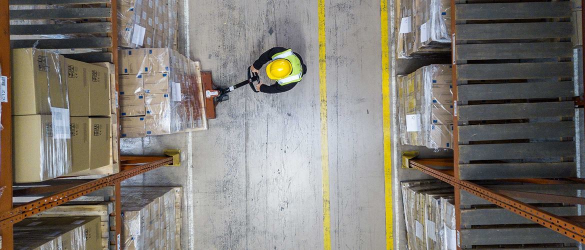 Mann mit gelber Warnweste und Schutzhelm bewegt Palette im Lager in orangenes Regal. Vogelperspektive