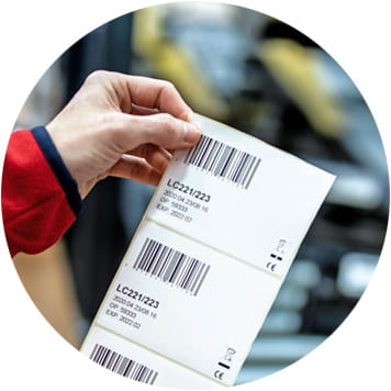 Logistikmitarbeiterin in rotem Pullover hält mehrere Barcode-Etiketten, Transportband mit etikettierten Päckchen im Hintergrund