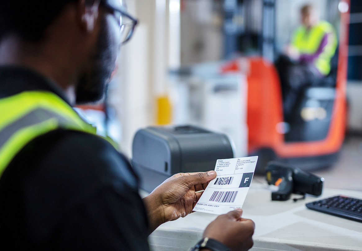 Logistikmitarbeit ein Arbeitssplatz Sitzend, hält Gedrucktes Brother-Versandetikett in der Hand, gapelstapler im Hintergrund