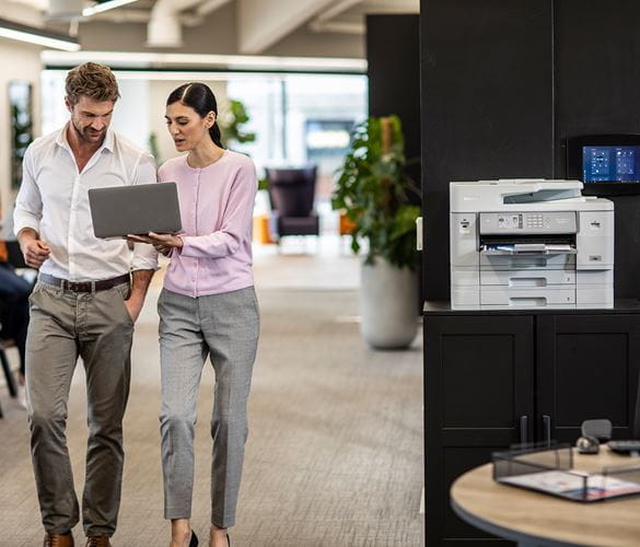 Zwei Personen laufen mit einem Laptop durch ein Büro und unterhalten sich. Tintenstrahldrucker von Brother im Hintergrund.