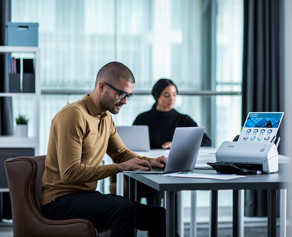 Ein Mann sitzt am Tisch und scannt ein Dokument ein. Frau im Hintergrund am Laptop.
