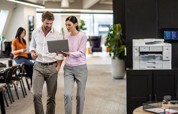 Eine Frau und ein Mann laufen mit einem Laptop durch das Büro. Im Hintergrund das Büro und ein Brother Drucker.