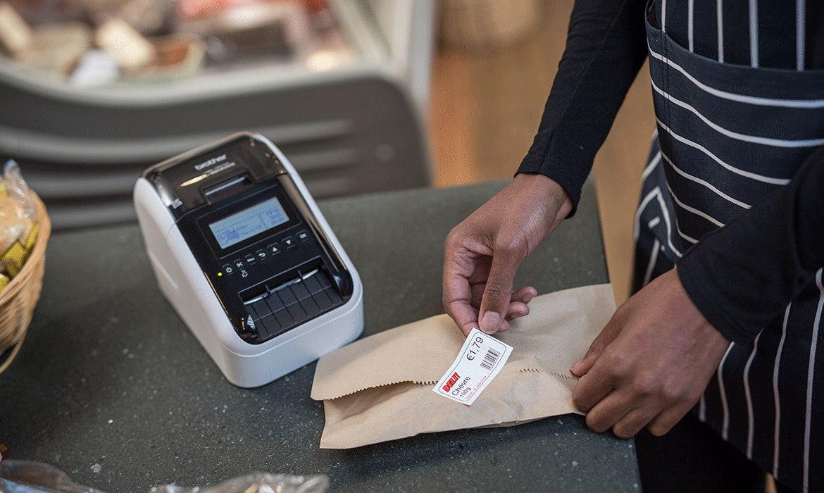 Eine Person klebt ein Preisetikett auf einen Umschlag, Brother Etikettendrucker für Preisetiketten im Hintergrund