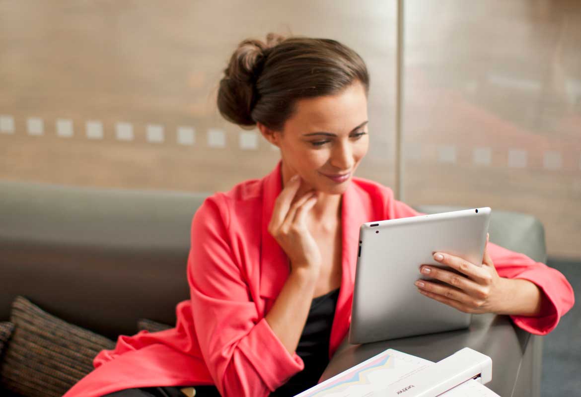 Einer Frau hält ein Apfel-Tablet in der Hand und Sitzt Neben einem Mobile Airprint-Drucker.