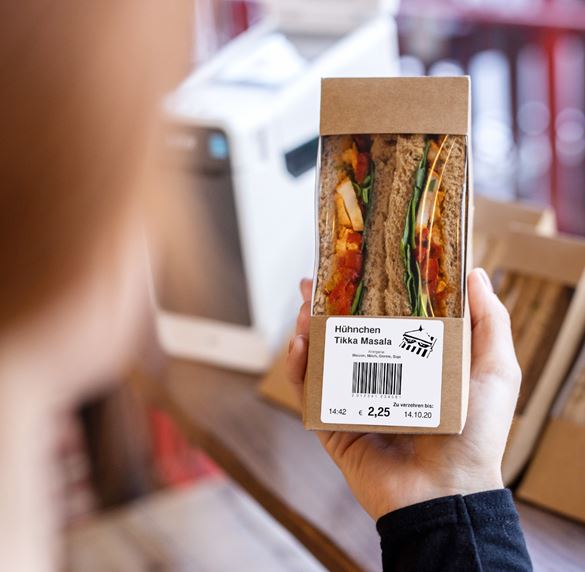 Eine Mitarbeiterin im Restaurant hält ein vorverpacktes Sandwich in der Hand, auf das Lebensmitteletiketten geklebt wurden