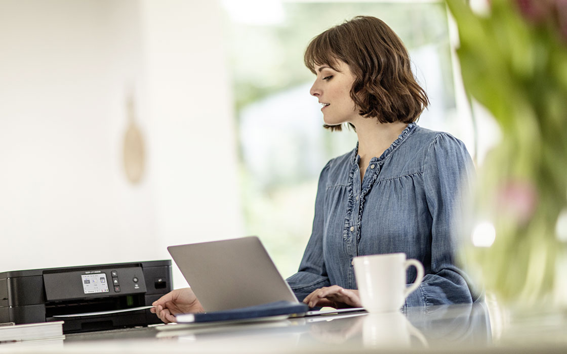 Eine Frau sitzt im Wohnraum am Laptop und Nimm, ein Seite aus dem Neben IHR Stehenden Schwarzen Wlan-Drucker.