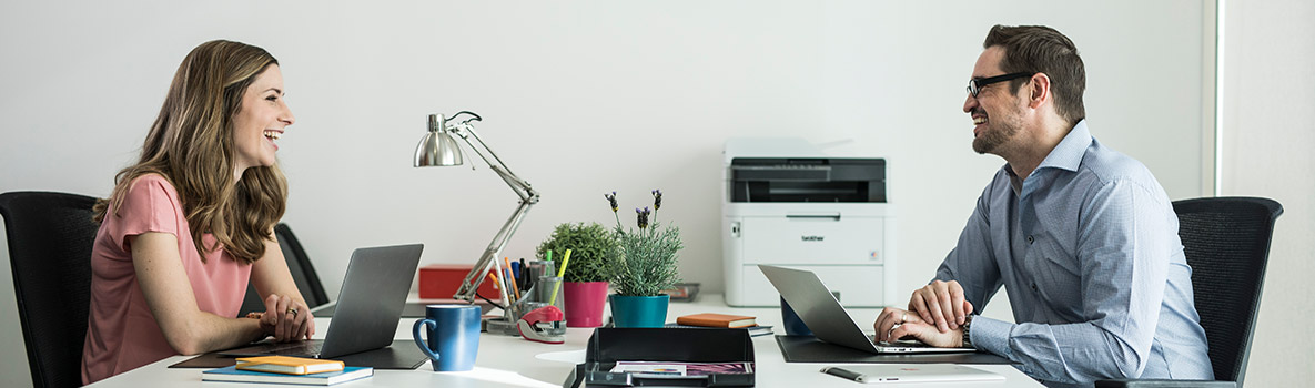Zwei Personen sitzen im Büro an Ihren Schreibtischen gegenüber und unterhalten sich.