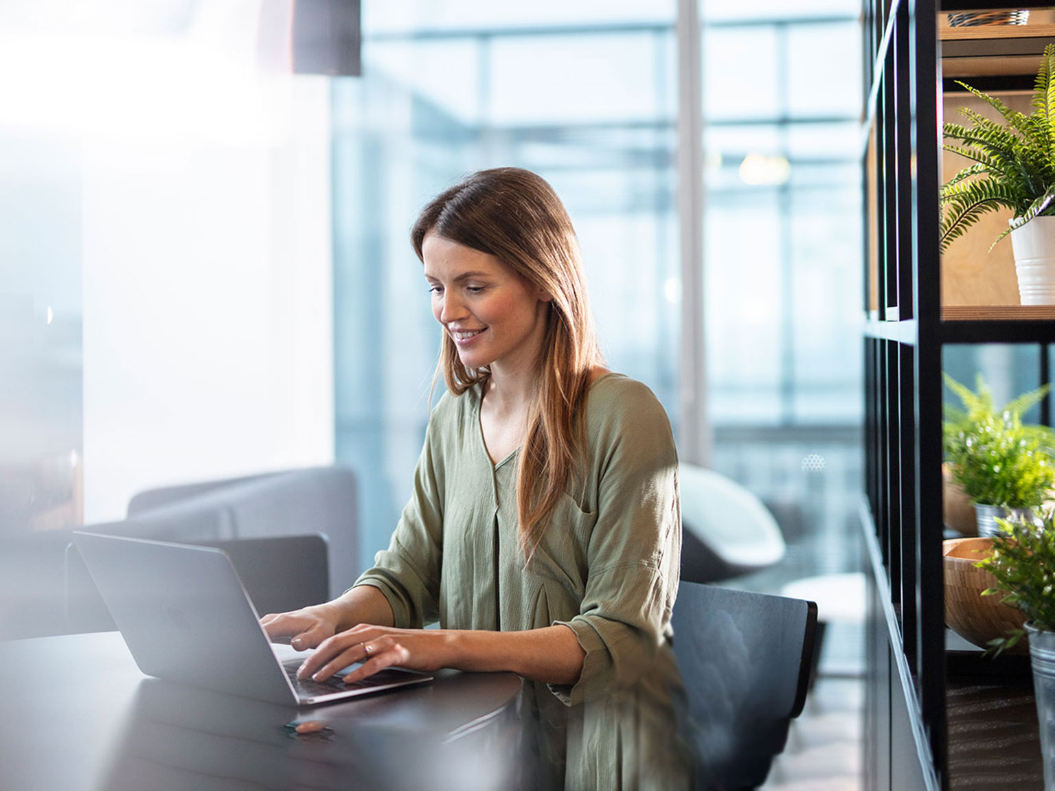 Woman sat at desk on a laptop sliming with a shelf in the background and green plants