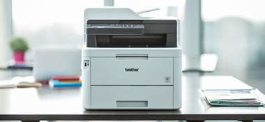 MFC-L3770CDW Multifunktionsfarben-Laserdrucker auf einem Schreibtisch mit Anlage im Hintergrund