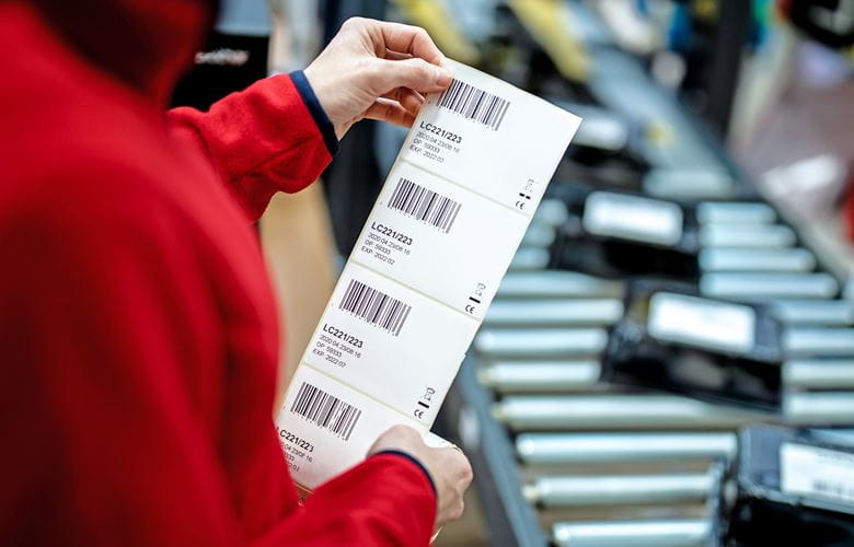 Logistikmitarbeitinin in Rotem Pullover Hält Mehreer Barcode-Etiketten, Transportband mit etikettierter Päckchen im Hintergrund