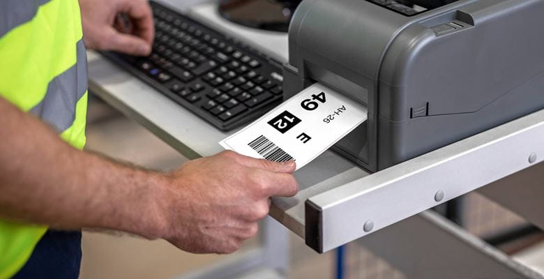 Logistikmitarbeit entnimmt Gerade Geedrucktes Regaletikett aus Brother TD-4T-Etikettendrucker, Tastatur und Bildschirm im Hintergrund
