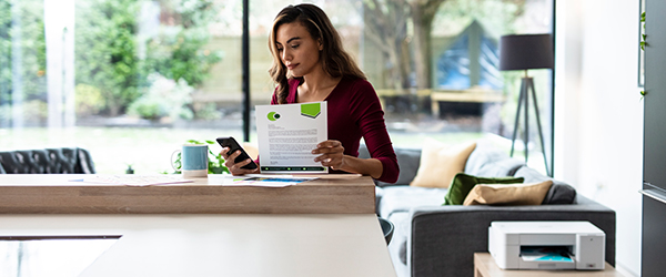 Frau im HomeOffice Hält Smartphone und Farbdokument in der Hand, Nebens IHR jeeht Einrucker.