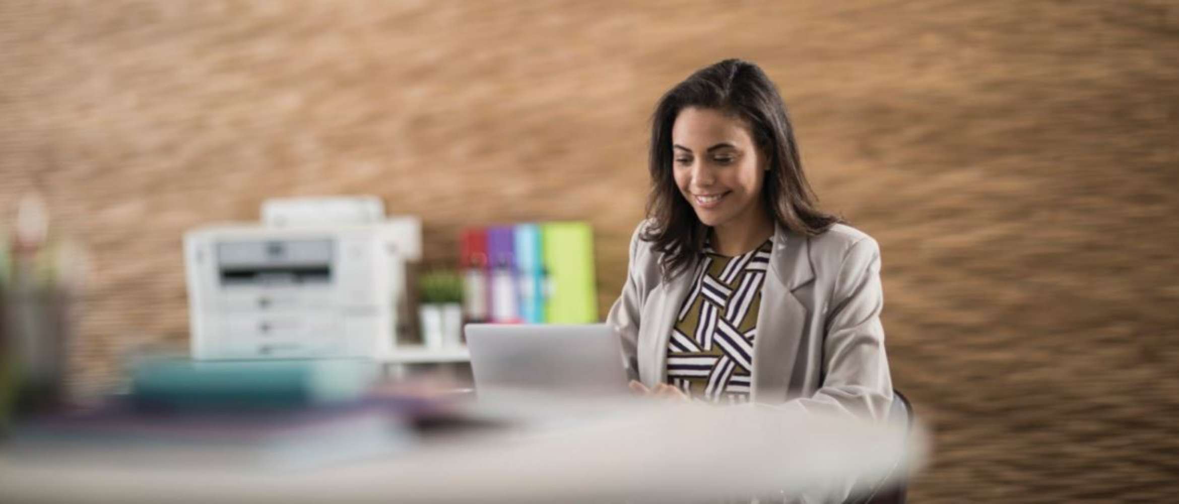 A female business worker in smart casual attire is sat working from a laptop a home office or small business scenario with a Brother multi-function SMB printer in the background