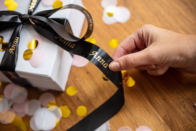 Hand holding black and gold personalised ribbon which is tied to a white box covered in yellow and white confetti