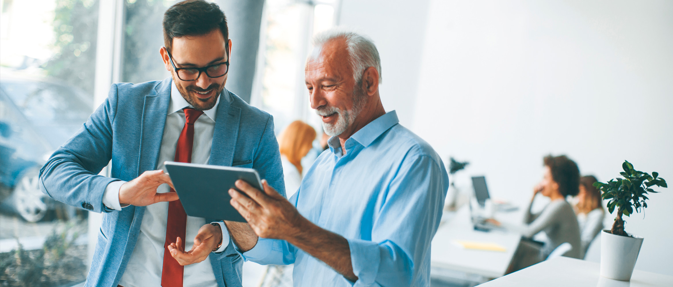 A young man in a bright suit and an older male co-worker in a more relaxed shirt discuss a business project on a tablet during a stand-up meeting in the more diverse and collaborative workplace of the future