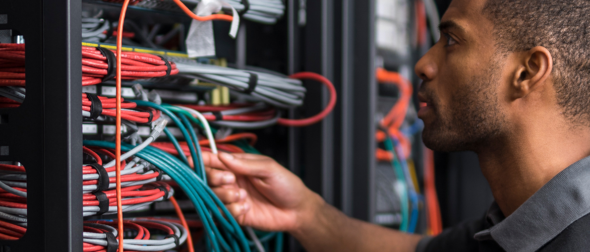 A young man in a grey polo shirt looking, with slight confusion, at groups of red, grey, and teal coloured network cables against a blurred background of more cables