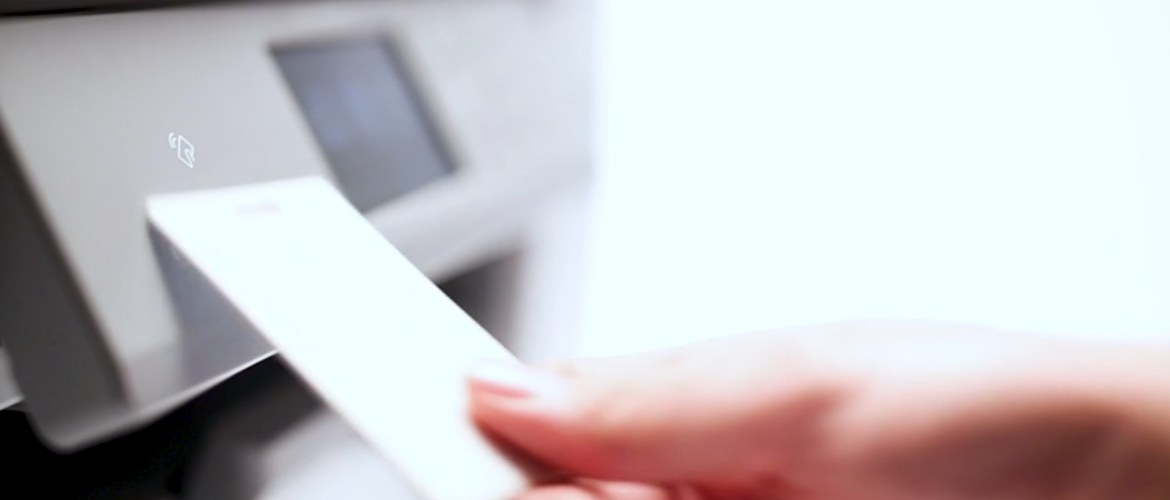 An office staff member taps an NFC (Near Field Communications) card against a Brother X-Series business inkjet printer to access the print functions and unlock secure printing. 