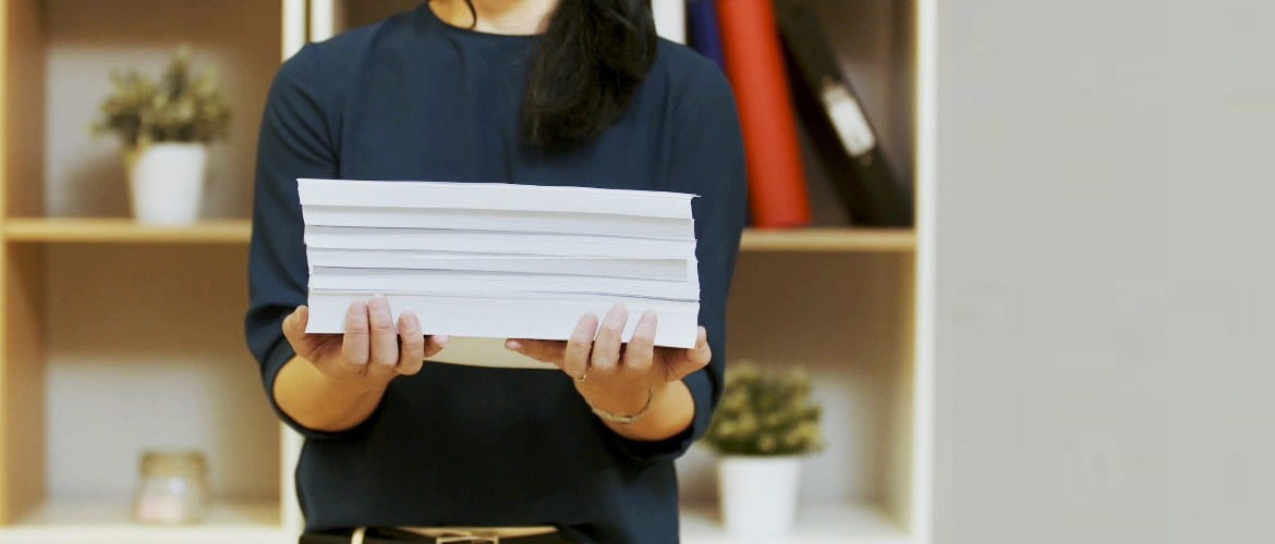 En kvinne i hjemmekontormiljø holder en bunke papirdokumenter som er skrevet ut