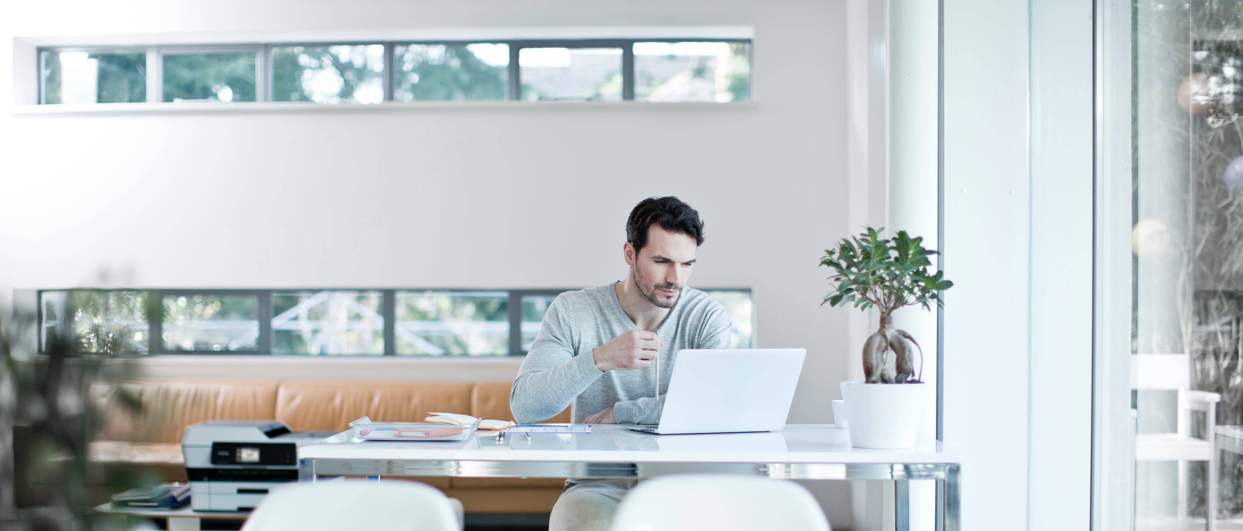 A man sitting at his desk in a home office set up in a bright room with two plants. A small printer is on the desk.