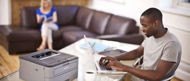 A man is sat in his home office at a desk working from a laptop computer with a Brother laser printer alongside him. In the background of the remote hybrid office scene is a woman sat on a brown sofa.