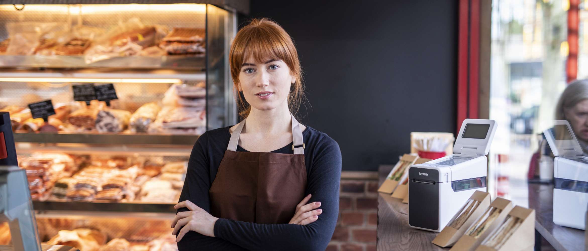 A female member of staff at a retail bakery store is standing arms folded looking to camera. A Brother label printer is in the background.