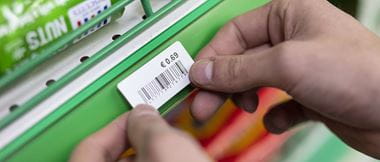 A staff member at a retail store is sticking a printed label onto a shelf edge. The label contains barcode information for scanning purchases. 