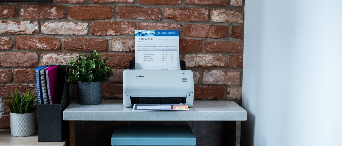 A Brother scanner sitting on top of a desk in front of an exposed brick wall with a printed paper in the output tray