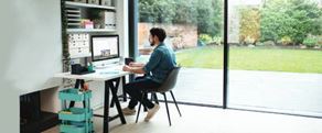 Nainen istuu työpöydän ääressä kotonaan katselemassa muistiinpanovälineitä ja kannettavaa tietokonetta. Etualalla on tulostin, kasveja ja tuoli.
