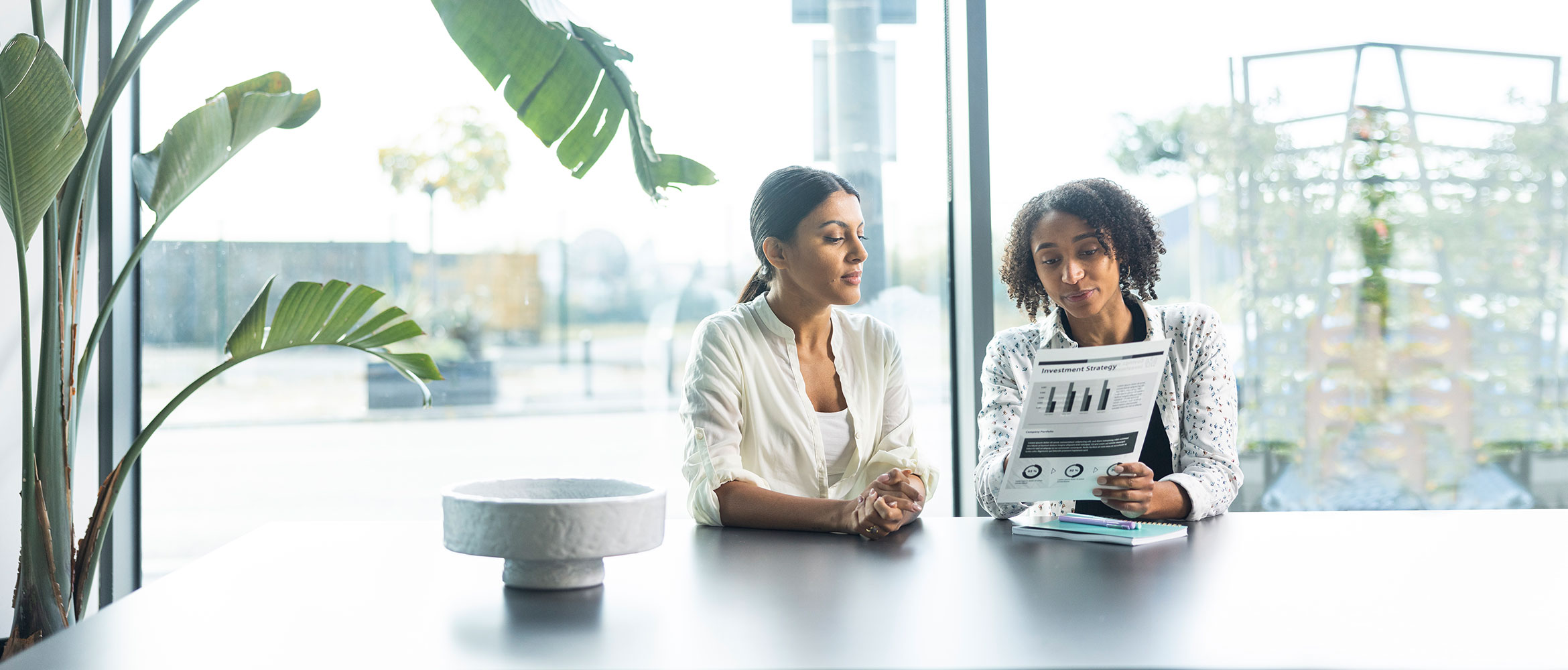 Man sat at table, woman in suit stood at printer in office, chairs, tables, plants, people in the background