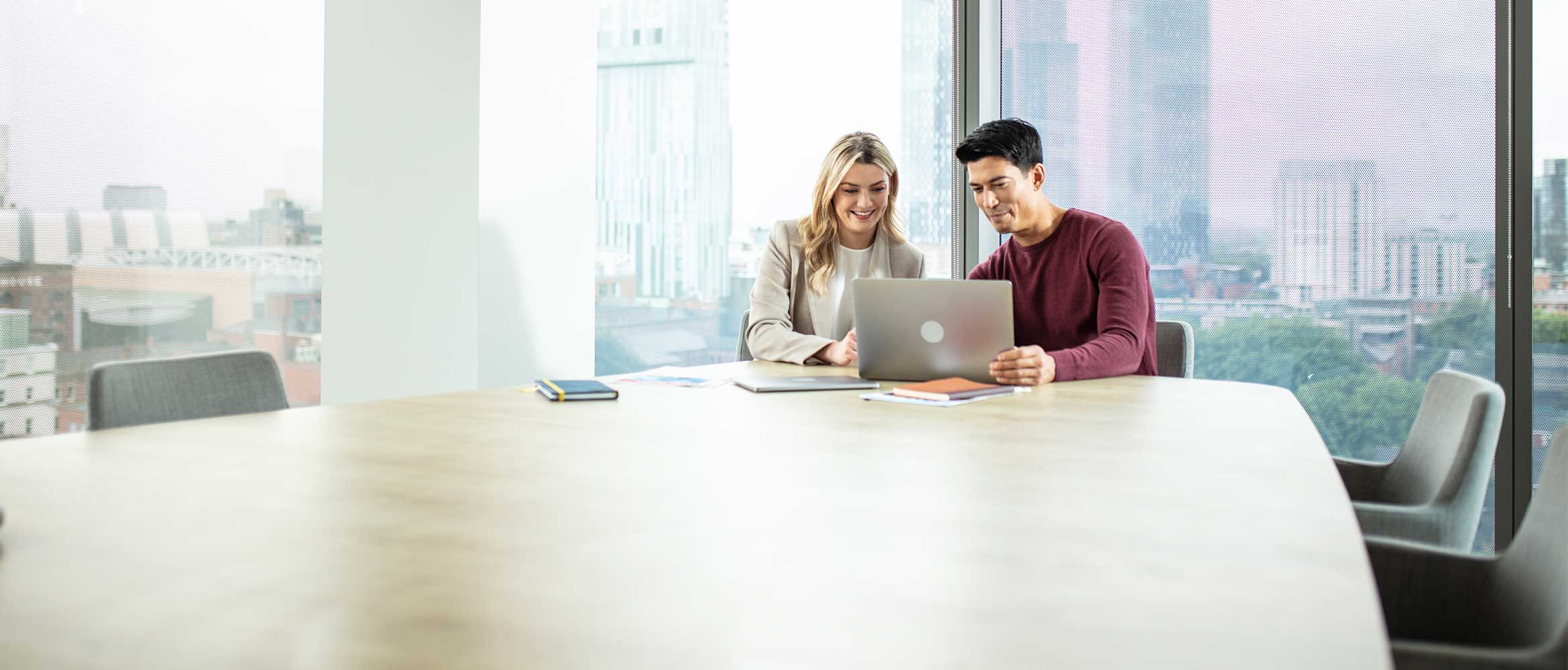 Man and woman sat around a desk look at laptop, window, chairs