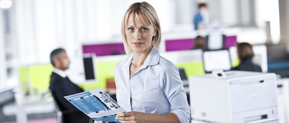 Žena drží výtisk ve velké kanceláři, v pozadí profesionální laserová tiskárna