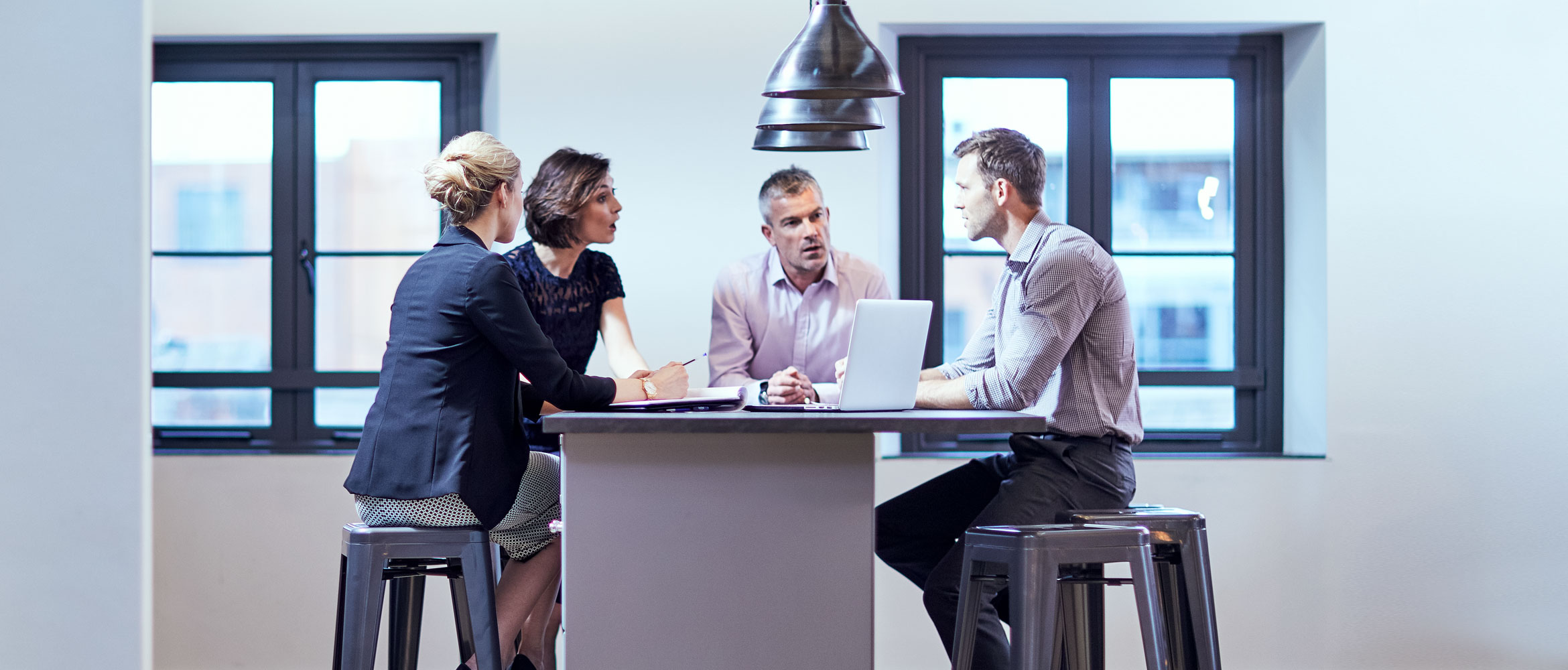 A group of four professionals sitting around a table, looking at a laptop screen and talking